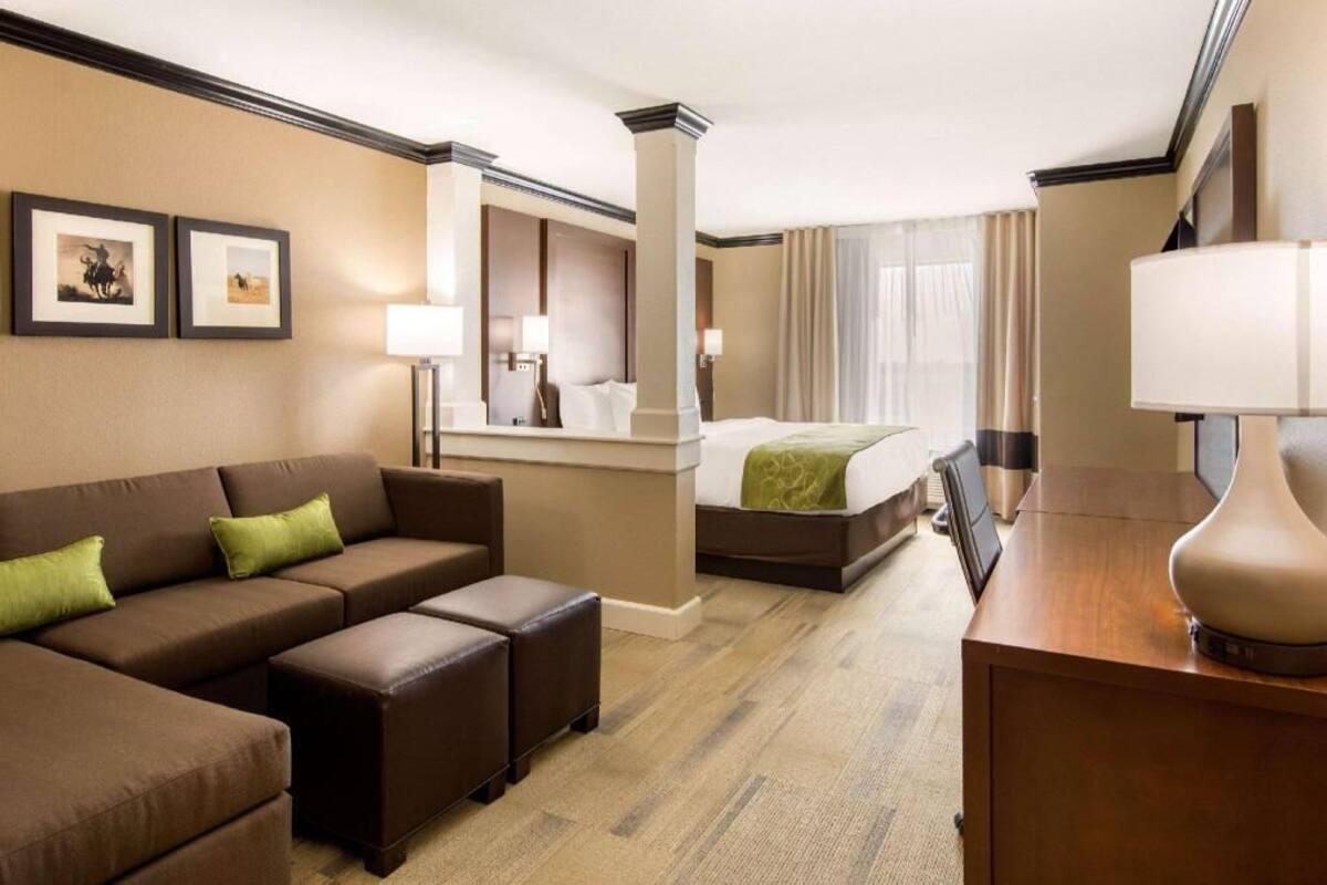 A hotel room with a sofa, ottoman, bed, bedside table, lamp, and a mirror.