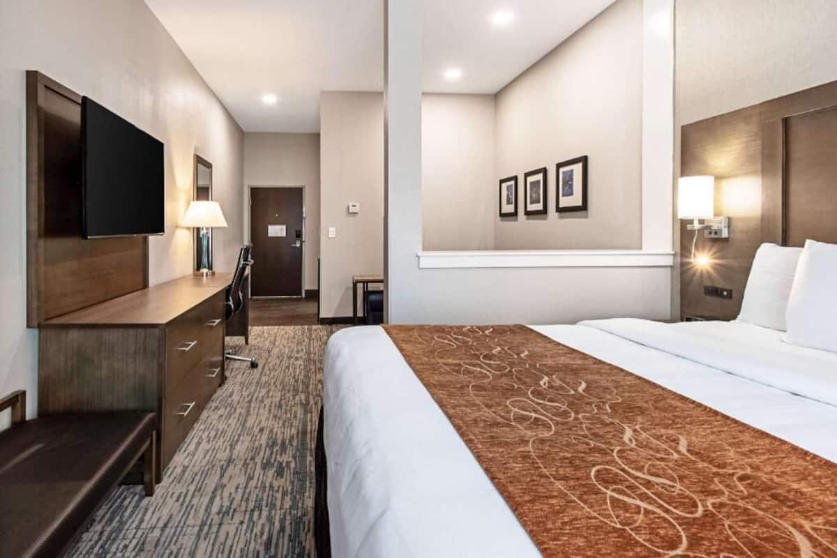 Quarto de hotel com cama, escrivaninha, cadeira, televisão e quadros nas paredes.
