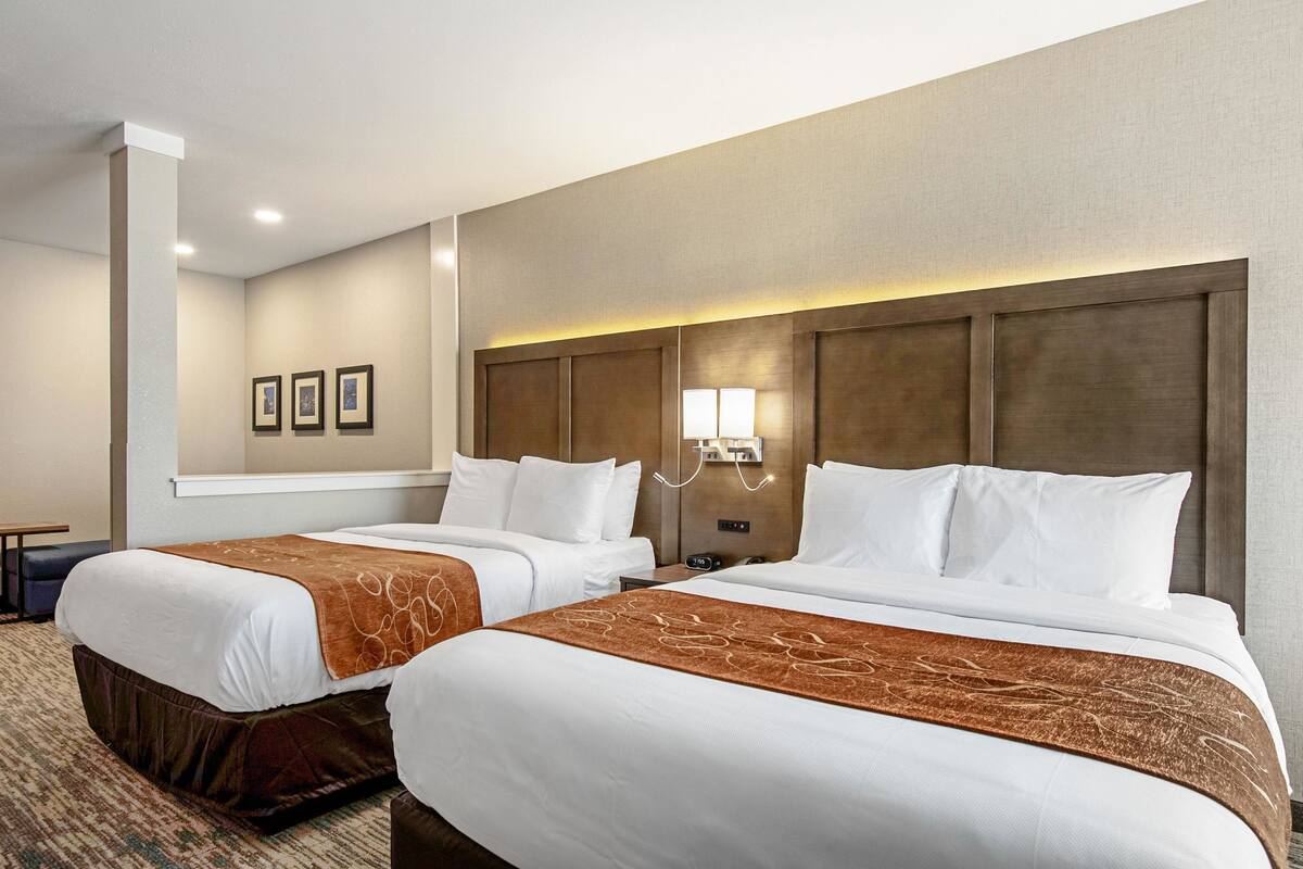 Quarto de hotel com duas camas, cabeceira com iluminação integrada, uma mesinha e um quadro pendurado na parede.