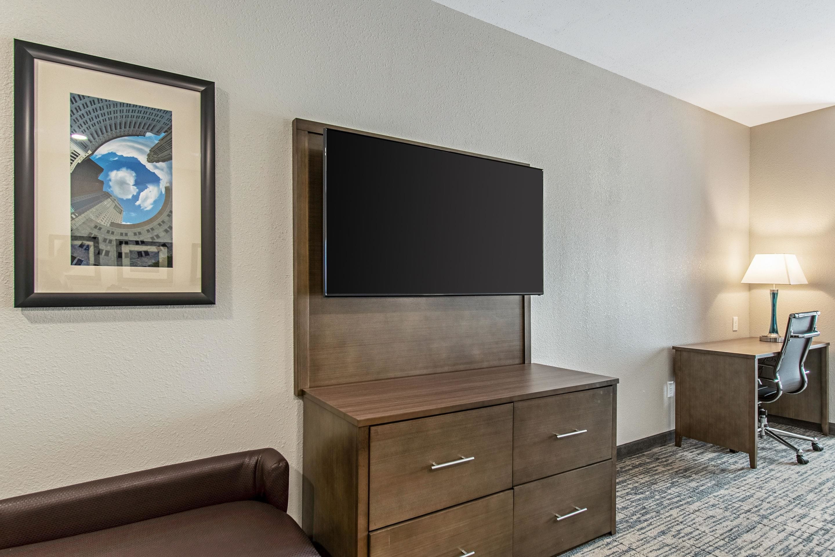 Quarto de hotel com televisão de tela plana presa a um armário de madeira, uma escrivaninha com cadeira e um quadro pendurado na parede.