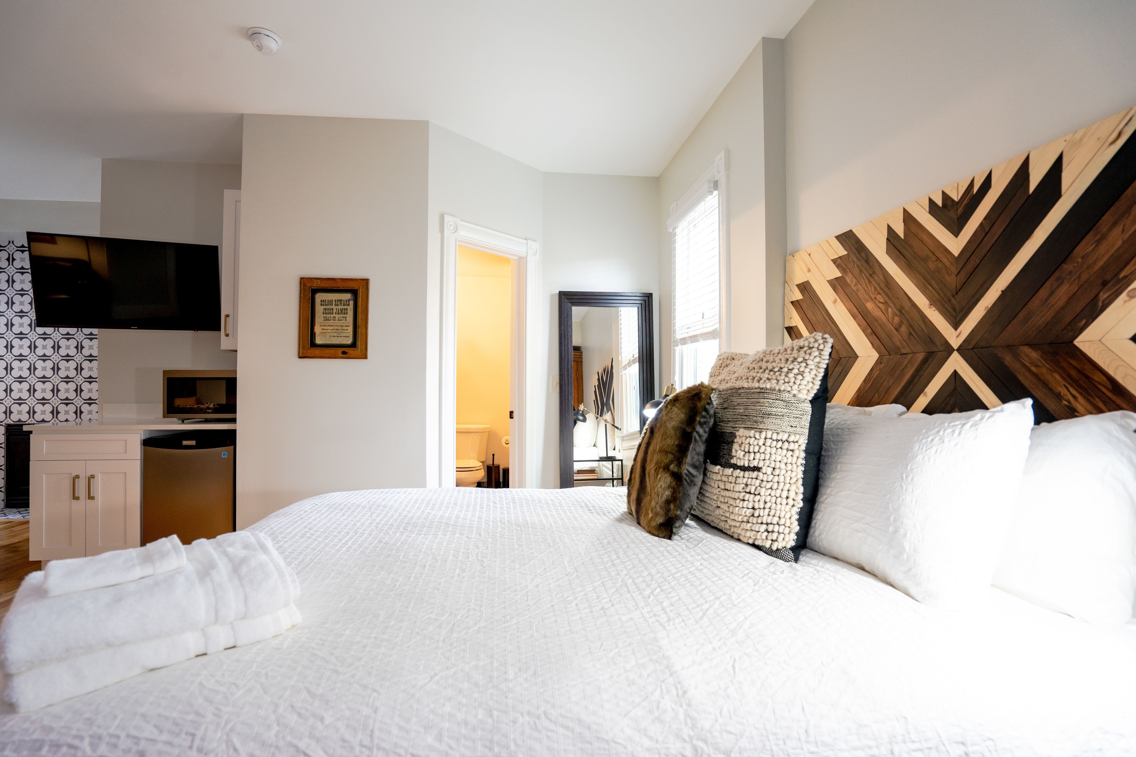Una camera d'albergo con un letto grande, una testiera in legno, una TV a schermo piatto, un angolo cottura e un bagno visibile attraverso una porta aperta.