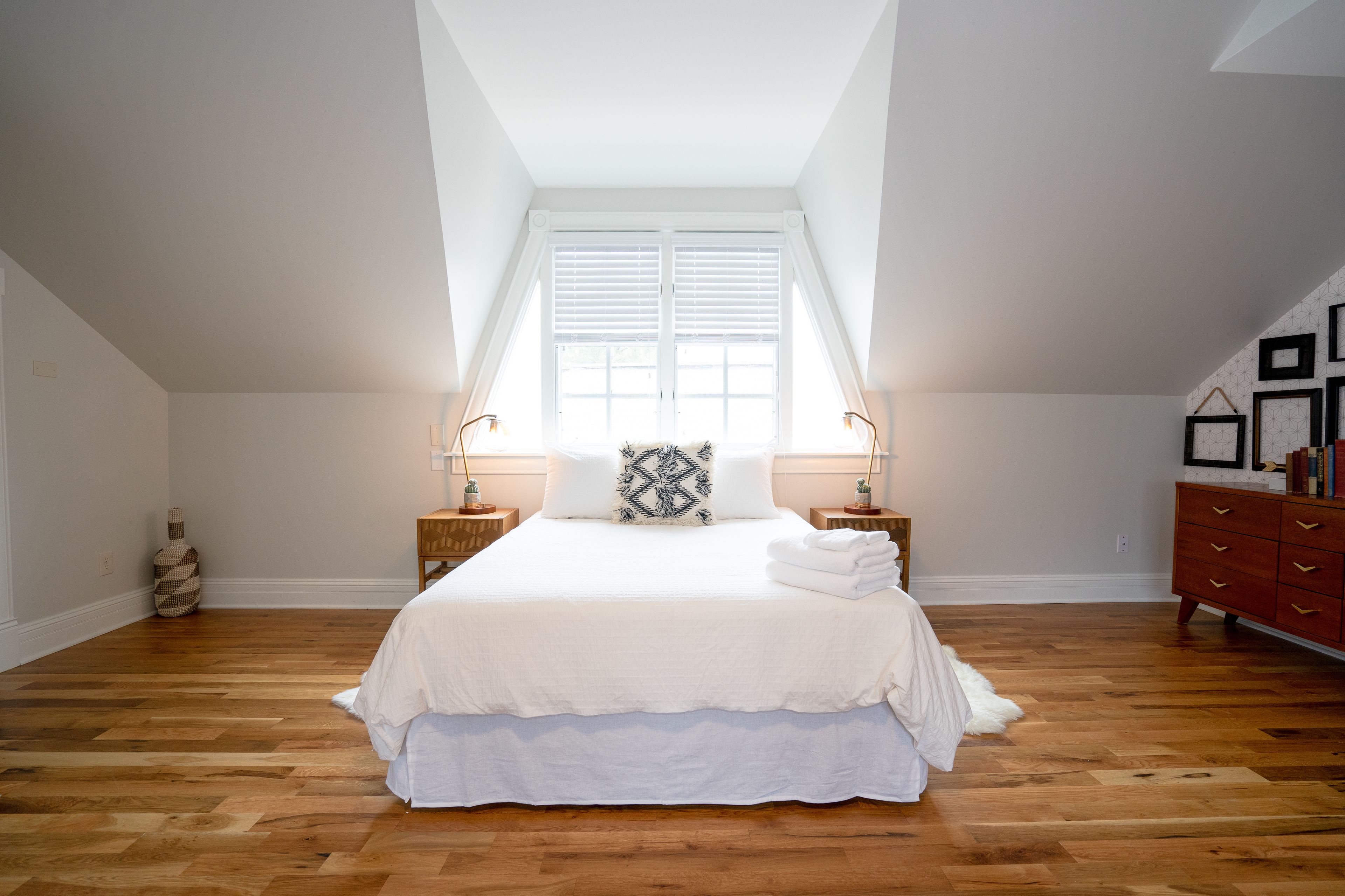 A bedroom with a large bed, a window with blinds, wooden flooring, a dresser, and two bedside lamps.