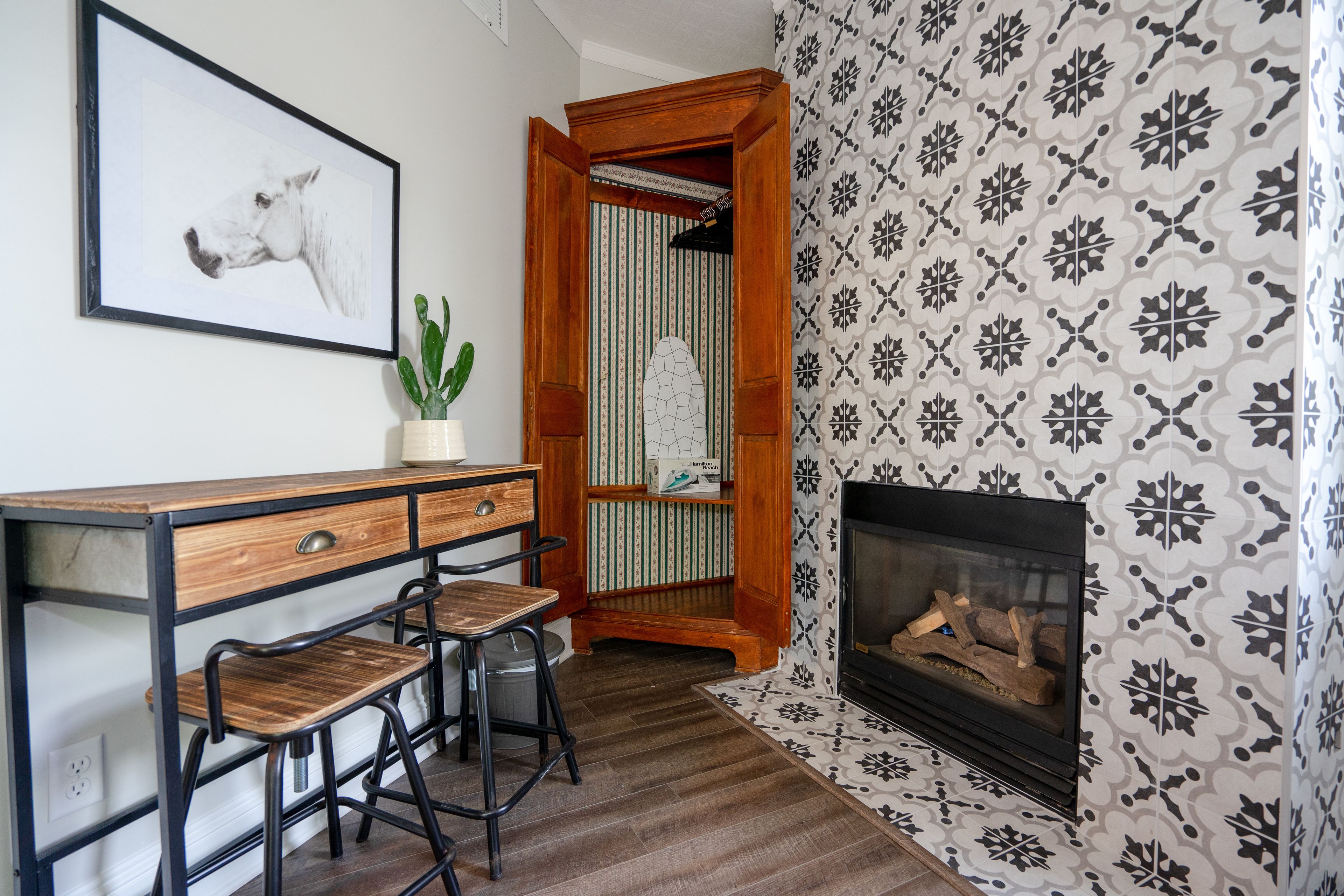 A room with a desk, two chairs, a framed picture, a potted plant, a patterned wall, and a fireplace.
