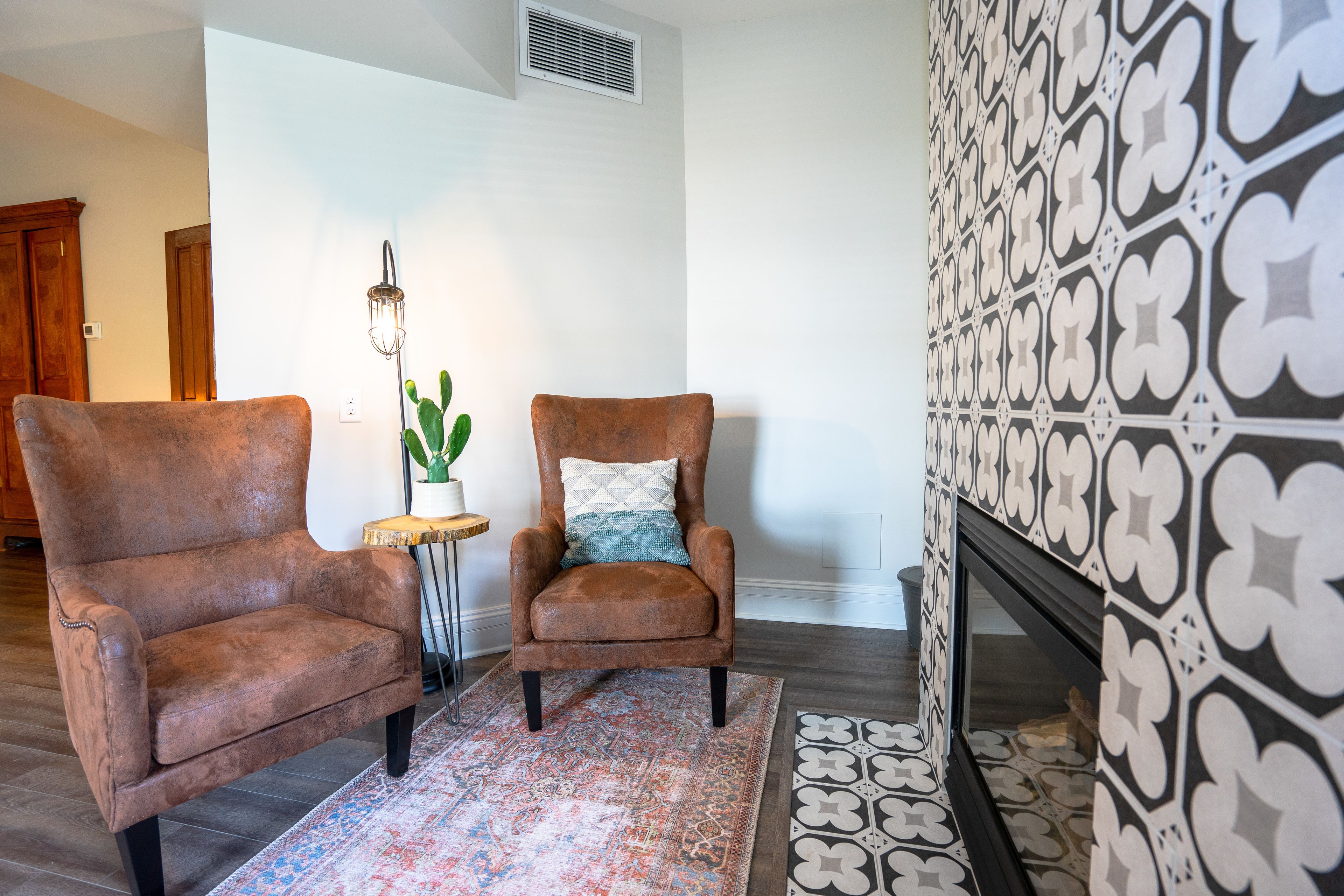 A room with two brown armchairs, a patterned rug, a small side table with a plant, and a wall with a geometric tile pattern.