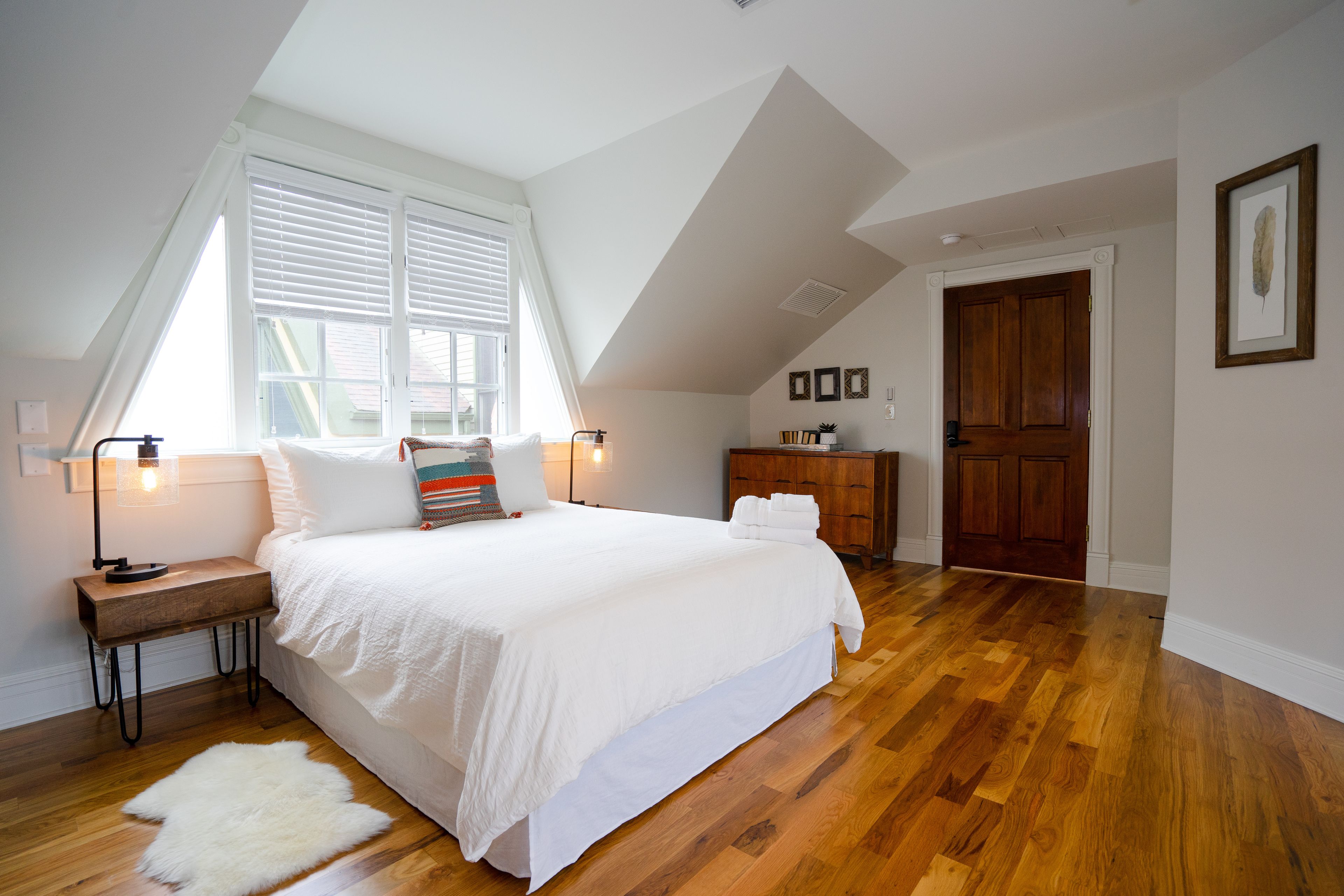 A bedroom with a large bed, wooden flooring, a window with blinds, a nightstand, a dresser, and a door.