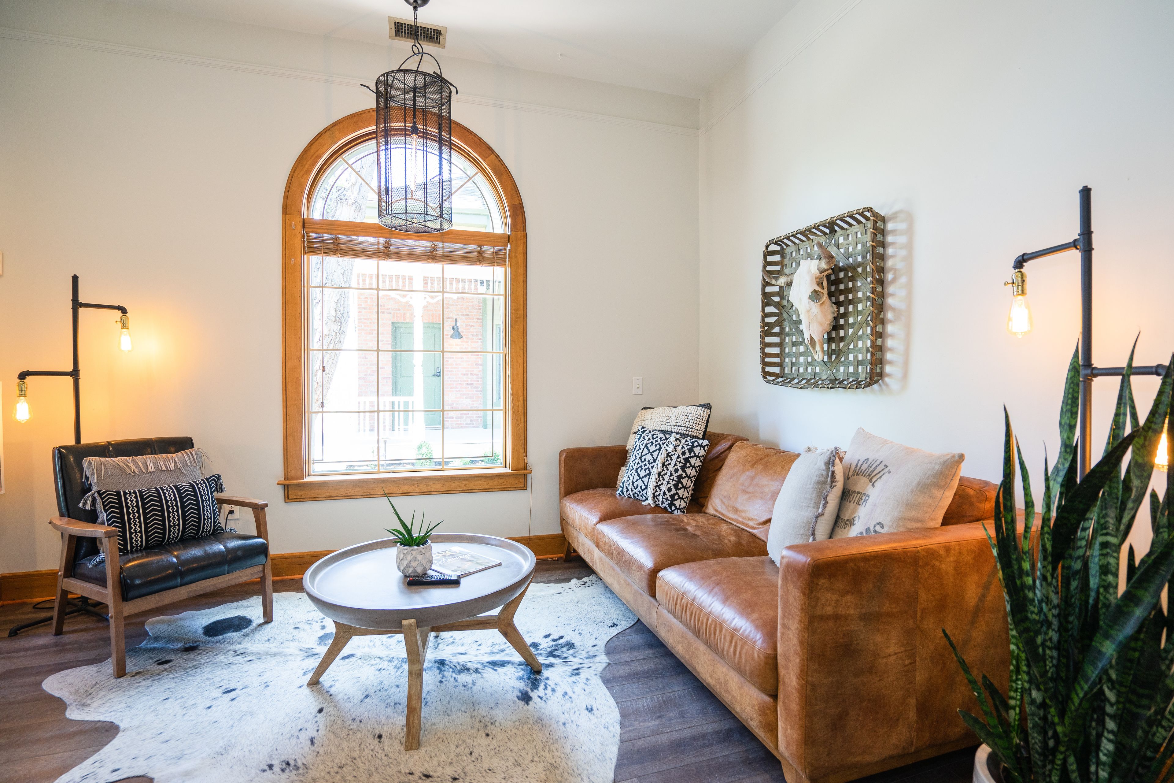 A cozy living room with a leather sofa, a wooden chair, a round coffee table, and a large window with a view of a brick building.
