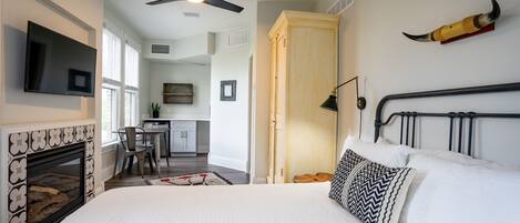 A bedroom with a bed, a fireplace, a television, a ceiling fan, and a kitchen area visible through an open door.