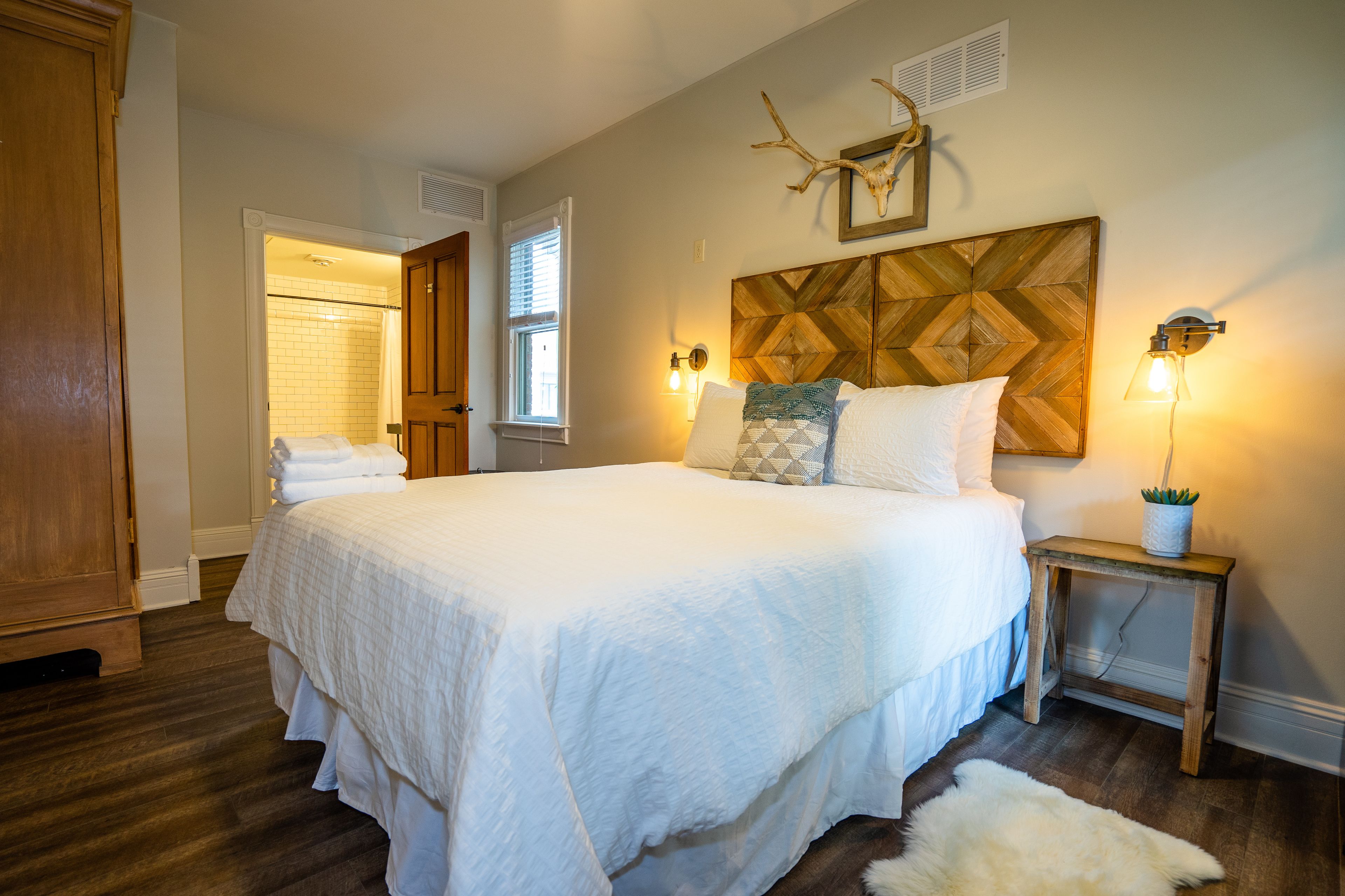 Ein Schlafzimmer mit einem großen Bett, einem Holzkopfende, zwei Nachttischlampen, einem kleinen Tisch mit Pflanze und einem flauschigen Teppich.