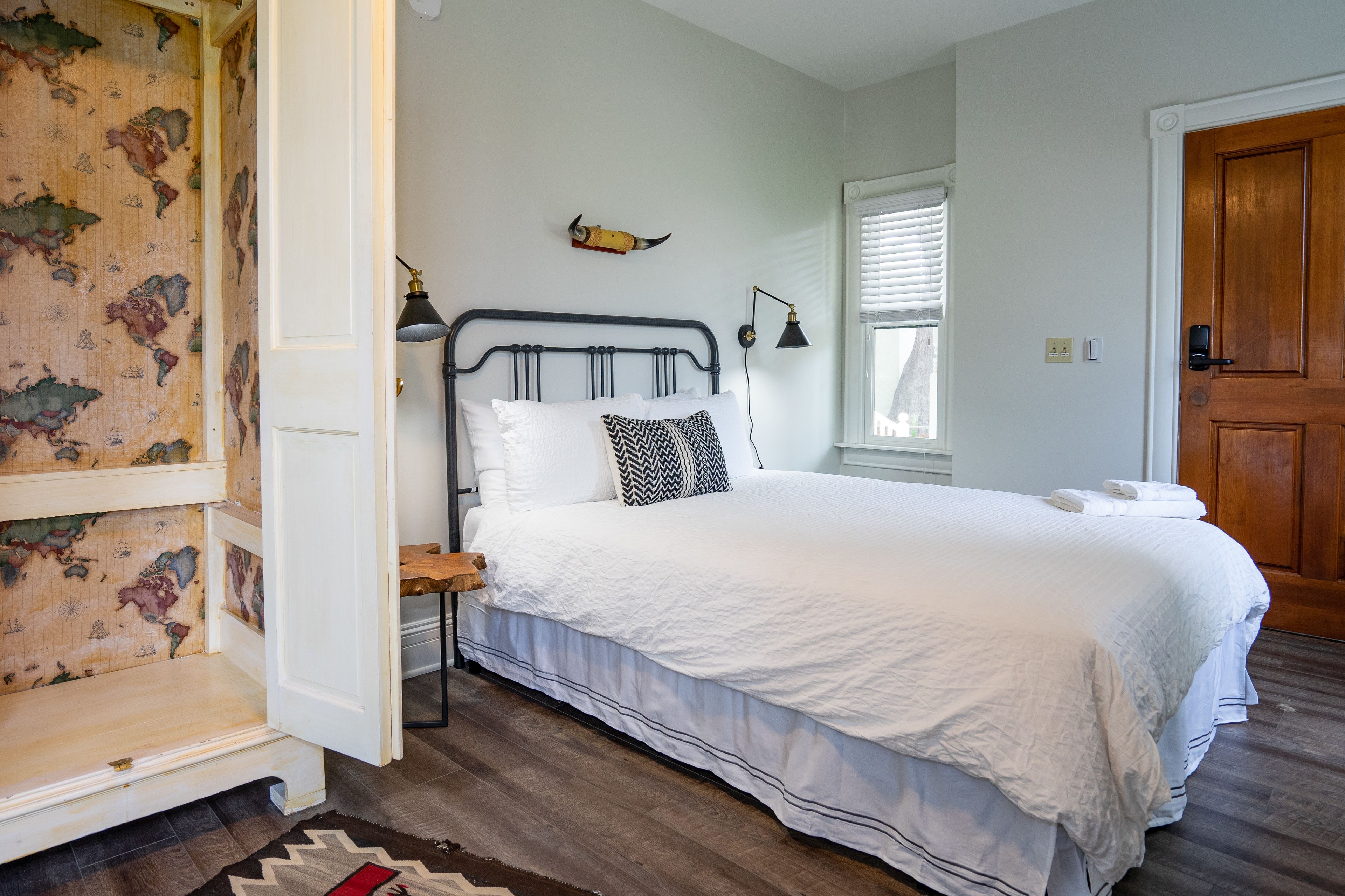 A bedroom with a bed, a nightstand, a lamp, a window with blinds, and a wooden door.
