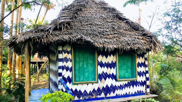 Una cabaña con techo de paja y exterior con diseño azul y blanco, rodeada de abundante vegetación.
