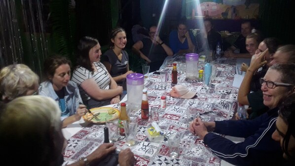A group of people seated around a table with a patterned tablecloth, various condiments, and a bottle of water. - Quarumanu Area (Tanna Island)