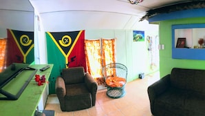 Una habitación con dos sillones, una mesita con un control remoto y un televisor. Hay dos banderas en la pared y una ventana con cortinas.
