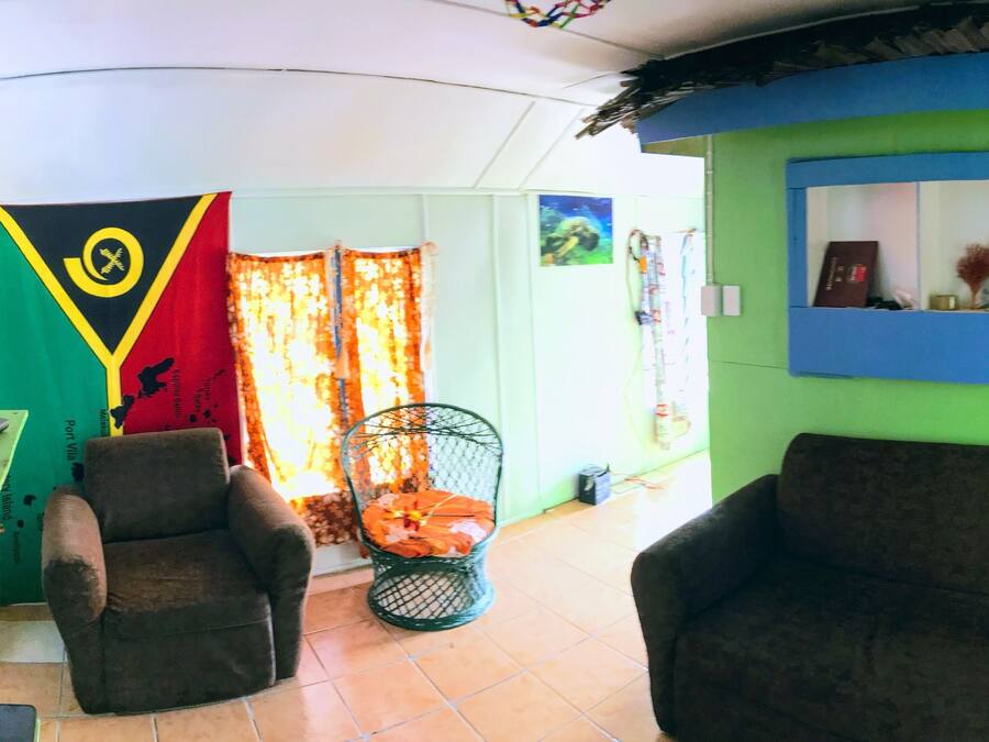 A room with two armchairs, a small table with a remote control, and a television. There are two flags on the wall and a window with curtains.