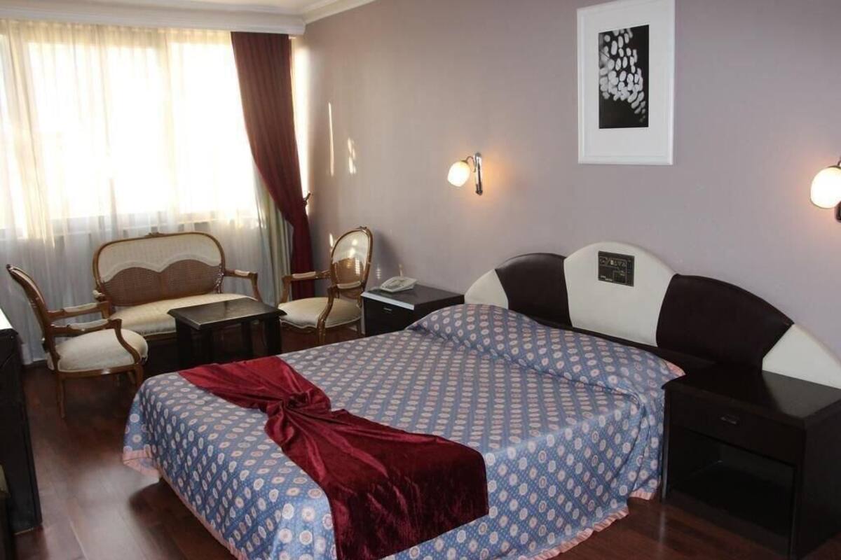 A hotel room with a bed, a desk, a chair, a nightstand, and a wall-mounted lamp.