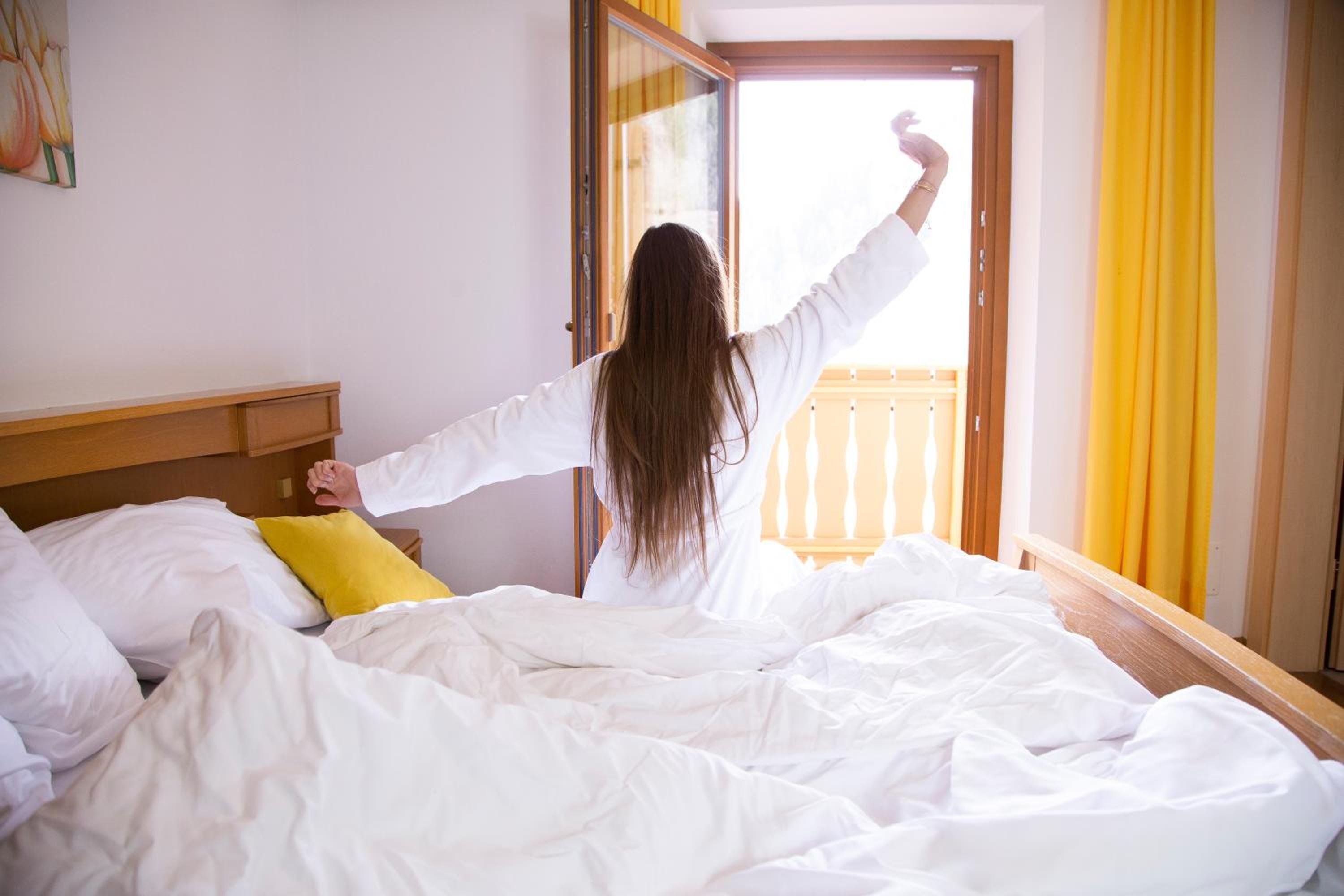 Quarto com cama, roupa de cama branca, travesseiro amarelo e cabeceira de madeira.