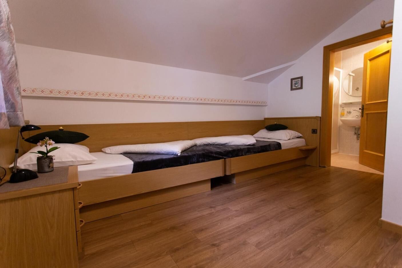 Ein Einzelbett mit Holz Kopfteil, ein Nachttisch mit Lampe und eine kleine Pflanze. Ein Badezimmer ist durch eine geöffnete Tür zu sehen.