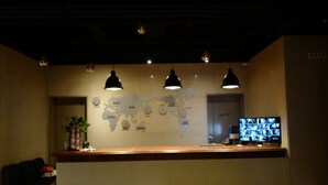 A reception area with a wooden counter, three pendant lights, a world map on the wall, a television, and a plant. - Renaissance Hotel - Hostel (Jeju City)