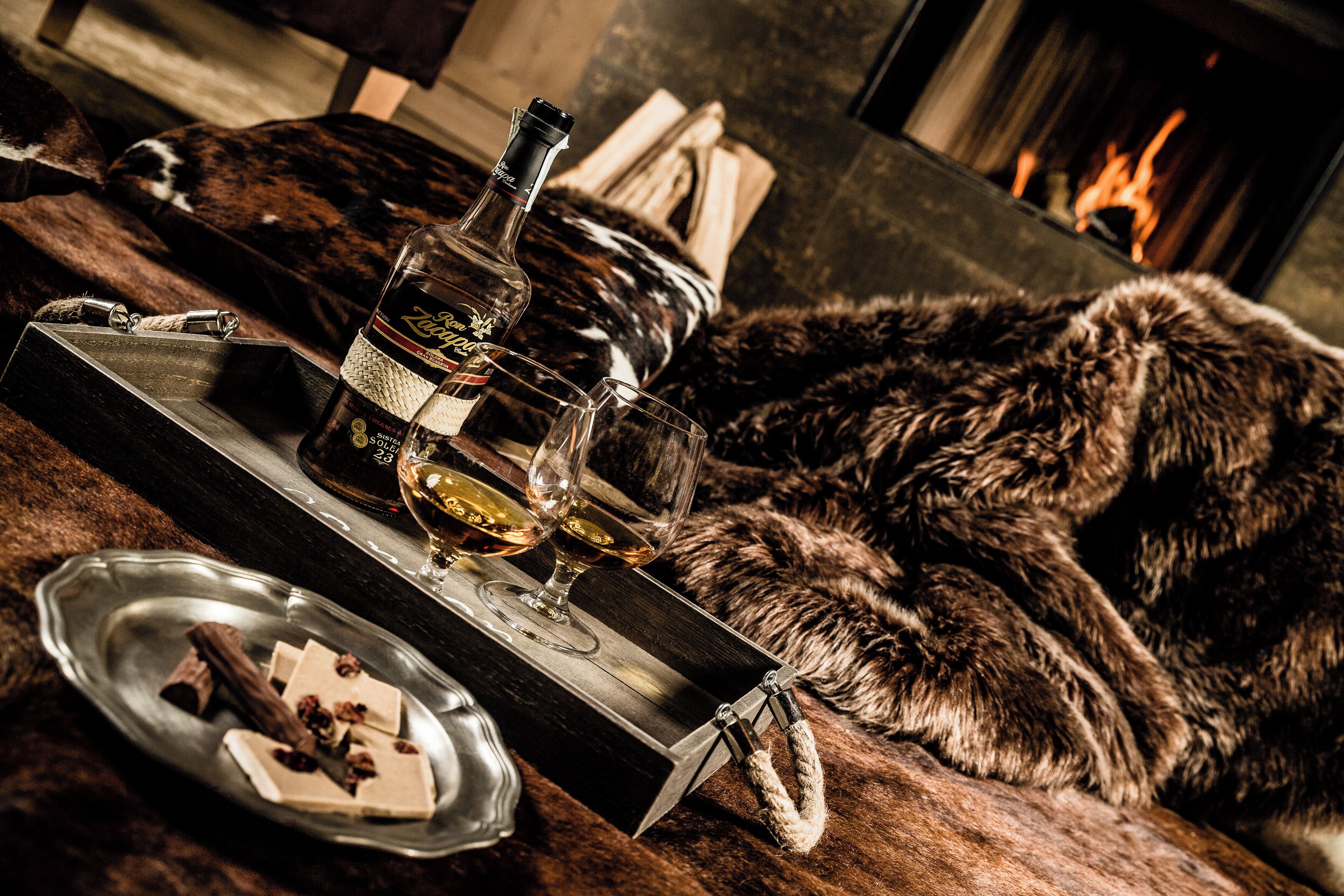 A cozy setting with a fur-covered couch, a tray with a bottle and glasses, and a plate with chocolate and nuts.