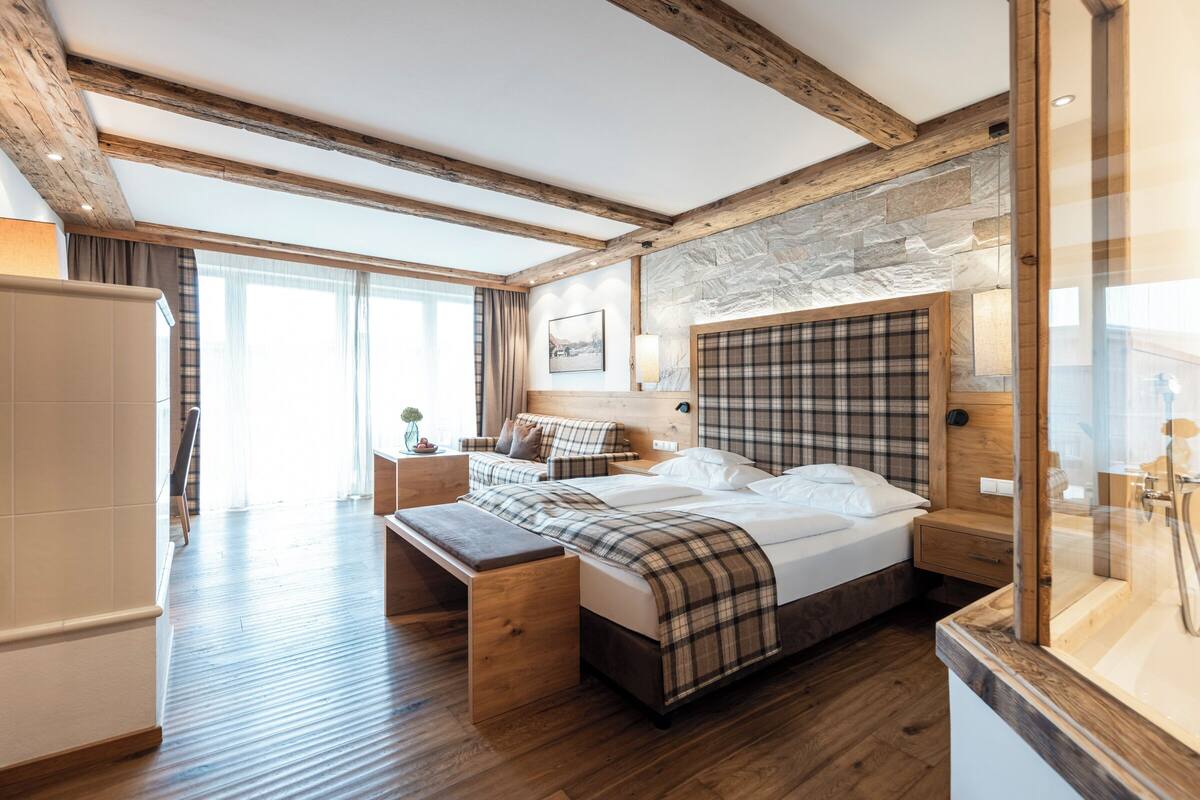 Ein modernes Hotelzimmer mit einem großen Bett, einer Bank, einem Holzbänkchen, einem Nachttisch und einem Badezimmer mit Spiegel und Waschbecken.