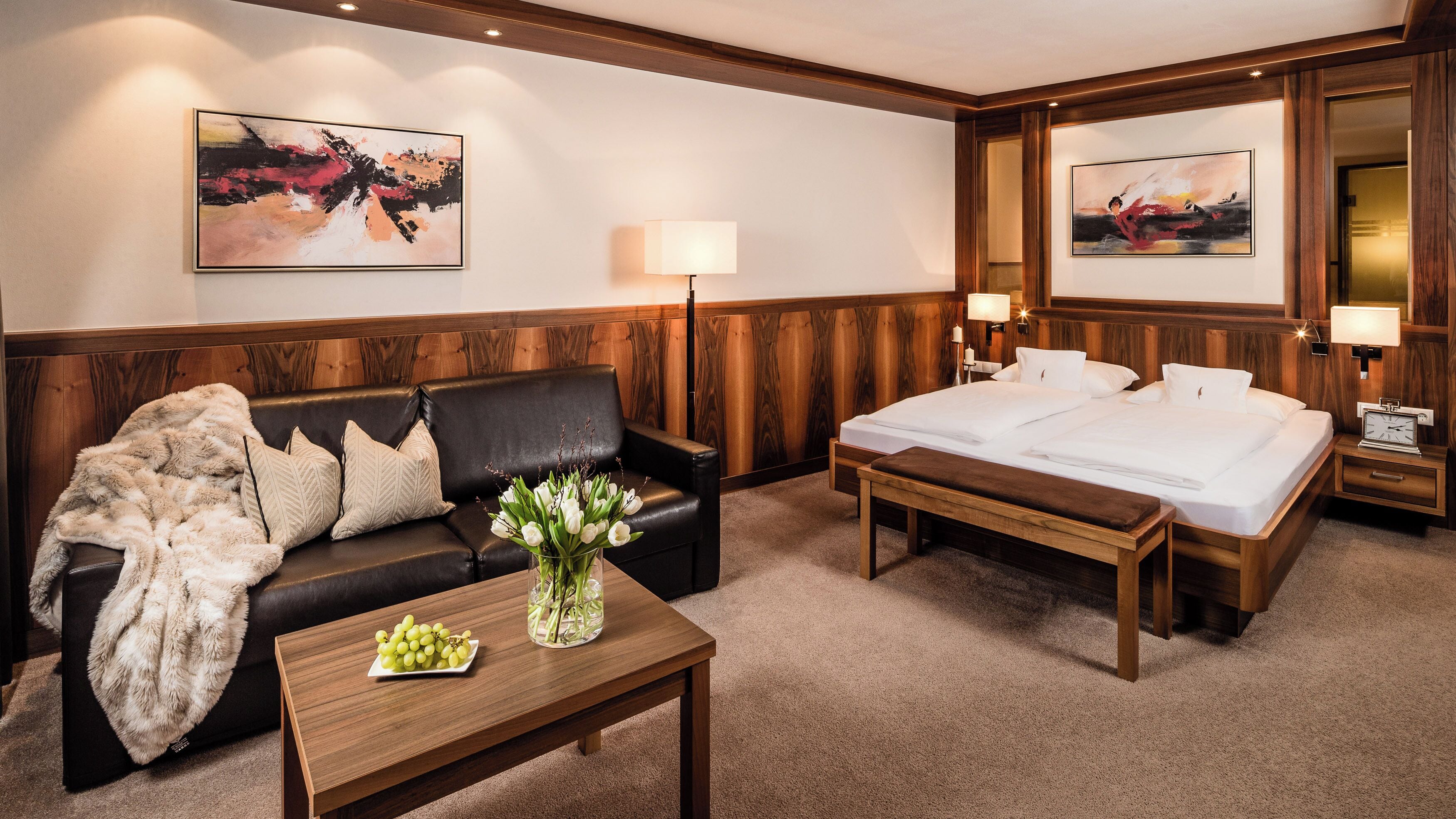 A hotel room with a wooden headboard, a black leather sofa, a wooden bed, a nightstand with a lamp, and a wall-mounted TV.