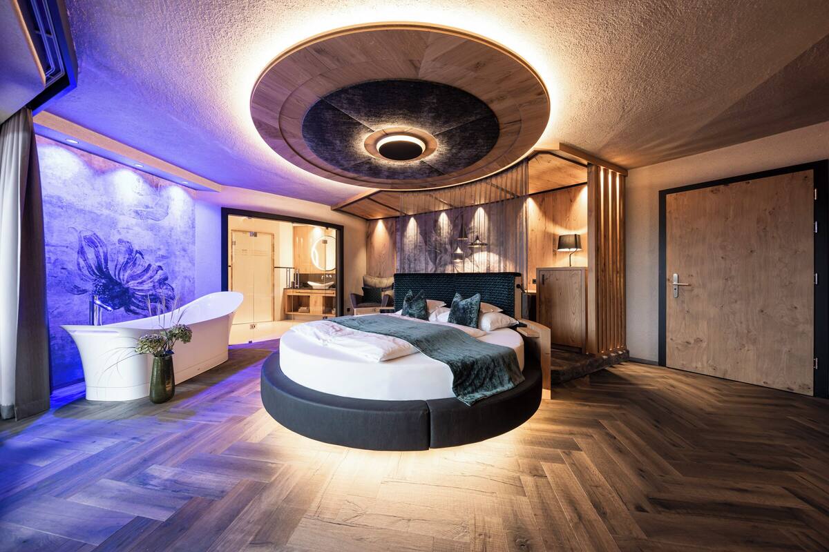 Ein rundes Bett mit Holz-Kopfteil, eine runde Deckenleuchte und ein großes Fenster mit Blick ins Freie.