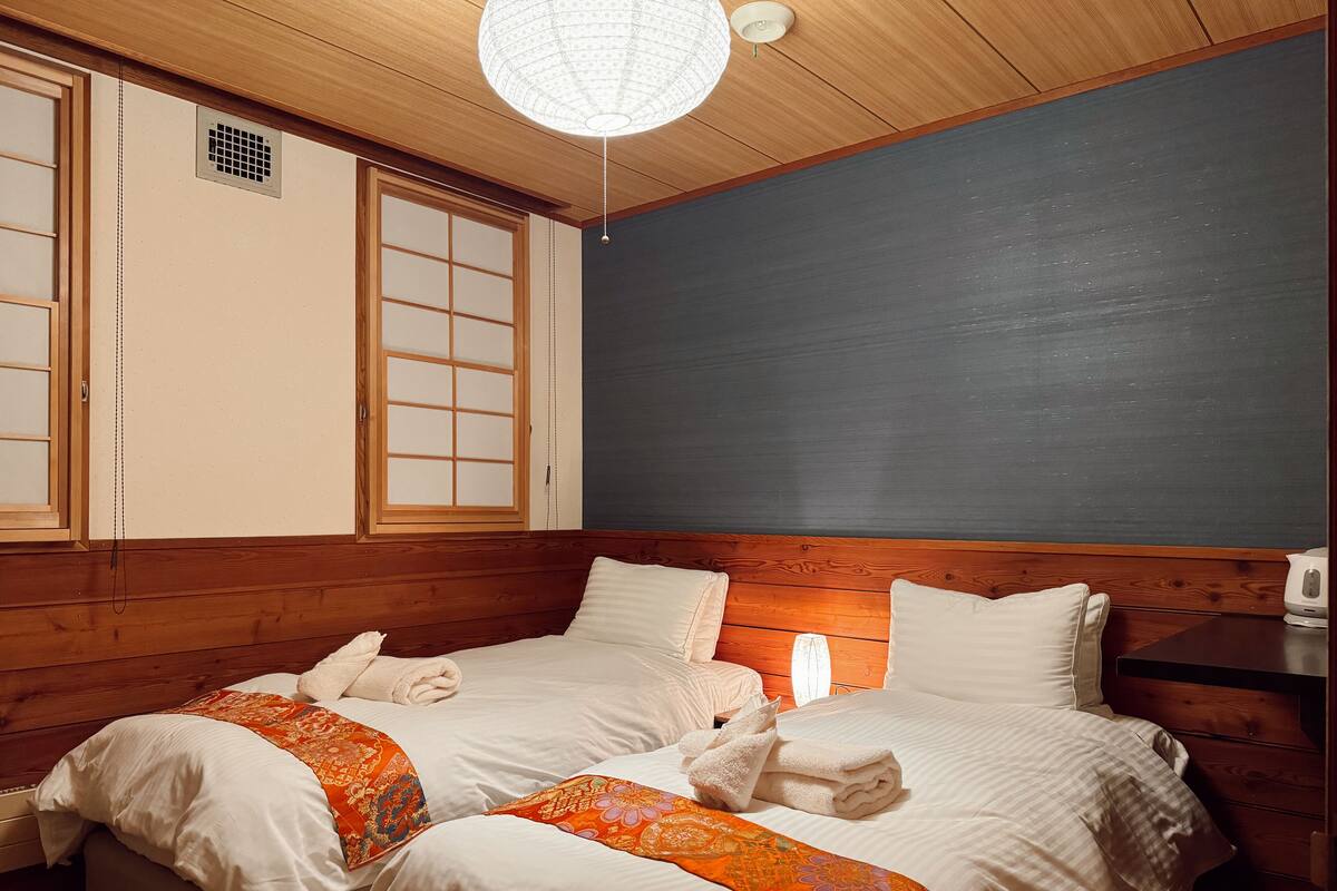 Quarto com duas camas, painéis de madeira, uma janela grande com biombos shoji e um lustre.