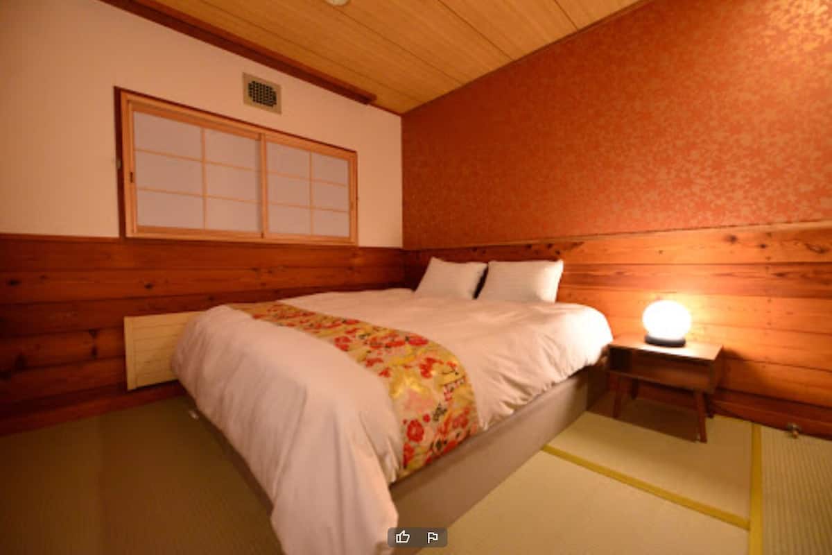 Quarto com cama de madeira, mesa de cabeceira com abajur e porta de tela shoji.
