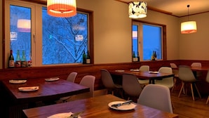 A restaurant with wooden tables and chairs, large windows with a view of trees, and pendant lights. - Raicho Lodge Madarao (Iiyama)
