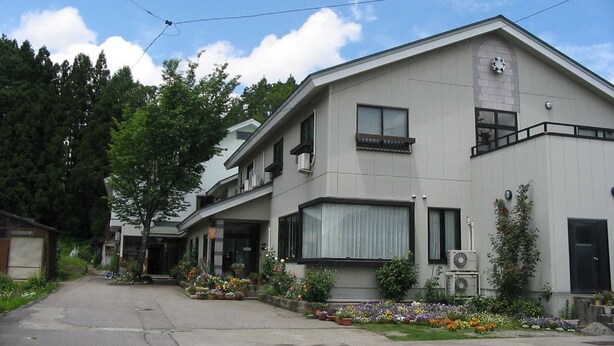 A two-story building with a flat roof, large windows, and a small balcony. There is a flower bed in front of the building. - Stayful House Nakamachi (Hakuba)