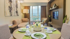 Habitación de hotel con una mesa de comedor preparada para dos personas, un sofá, una silla, un televisor y un amplio ventanal con vistas a la ciudad.
