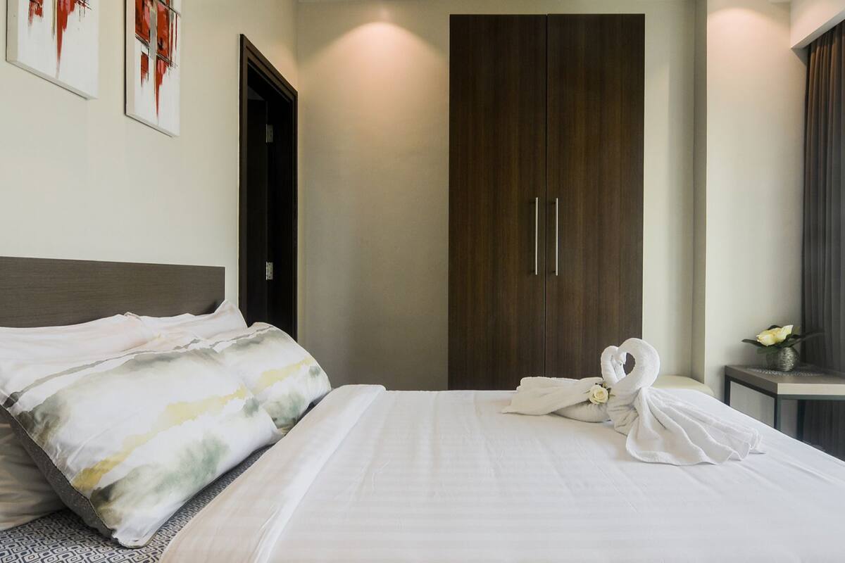 A neatly made bed with white linens, a dark wooden wardrobe, and a small bedside table with a vase of flowers.