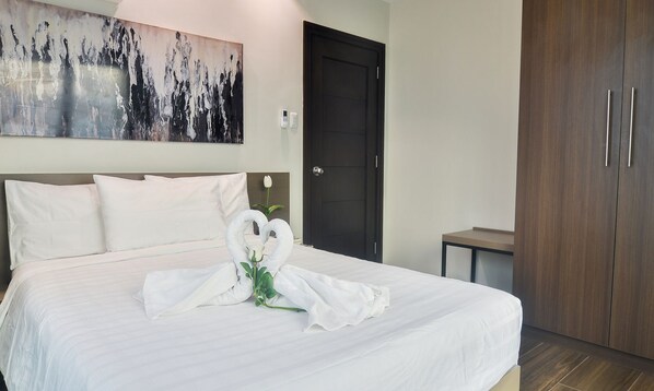 Una cama bien tendida con sábanas blancas, una toalla dispuesta en forma de corazón, una flor blanca y un cuadro abstracto grande en la pared.