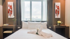 Habitación de hotel con una cama grande, ropa de cama blanca, un arreglo de toallas con forma de corazón, un jarrón con una flor y dos cuadros abstractos en las paredes.