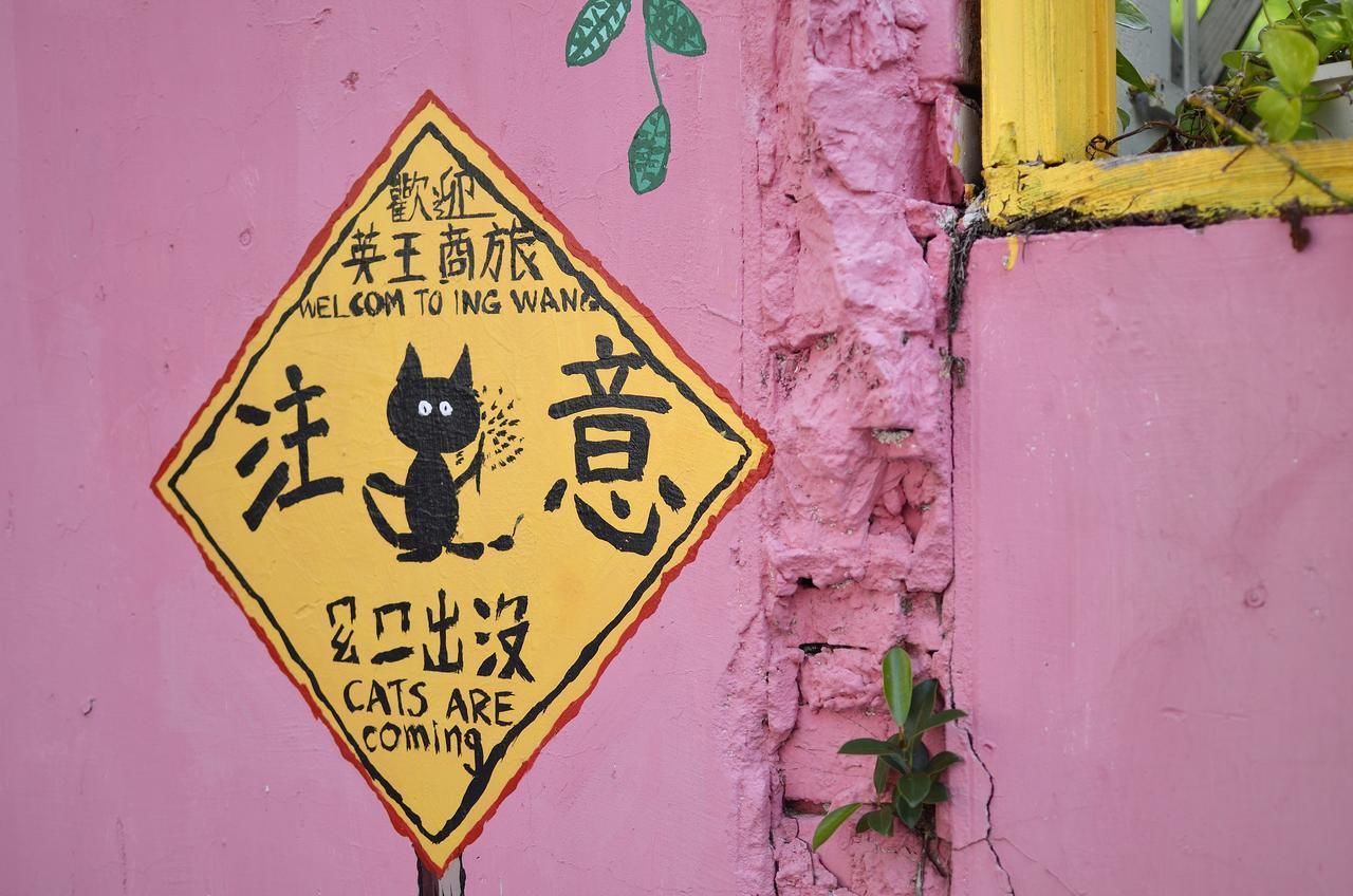 Una señal amarilla en forma de rombo con un gato negro y caracteres chinos, sujeta a una pared rosa.