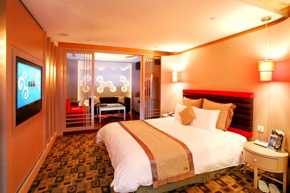 Quarto de hotel com uma cama grande, uma televisão na parede, uma mesinha de cabeceira com abajur e cabeceira vermelha.