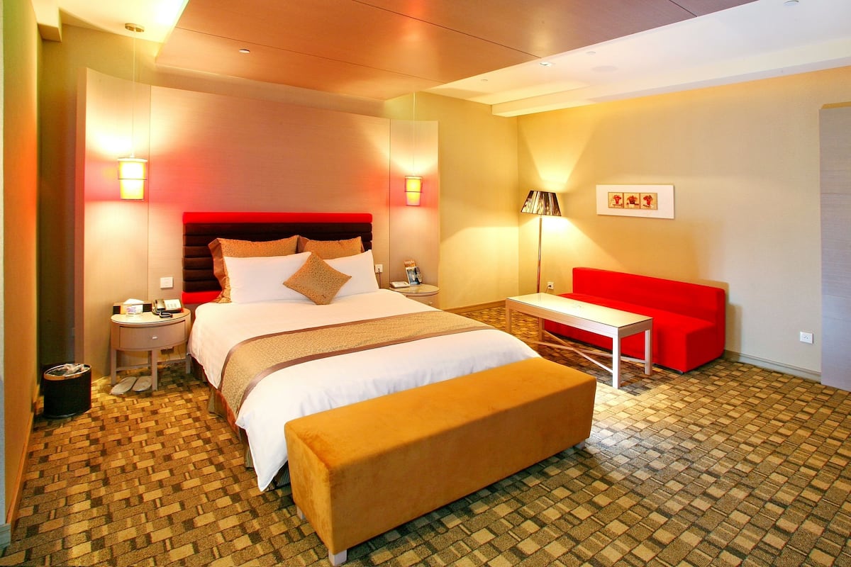 Quarto de hotel com uma cama grande, um sofá vermelho, uma mesa pequena e um banco.