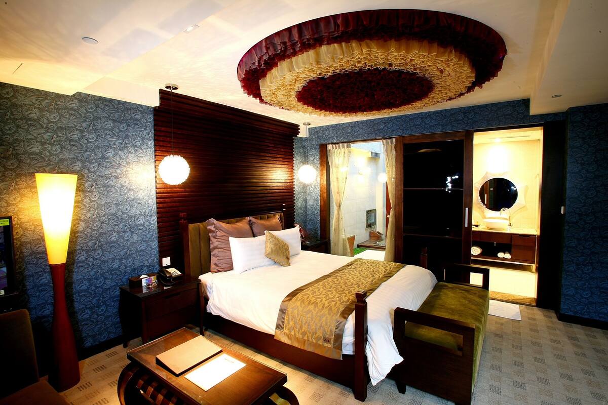 Quarto de hotel com uma cama grande, uma escrivaninha, uma cadeira, uma mesinha de cabeceira, um abajur e um espelho.