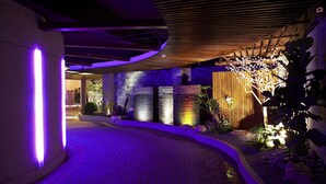 Una entrada moderna a un edificio con un camino de ladrillo curvo, iluminada con luces moradas, y una valla de madera con luces decorativas.