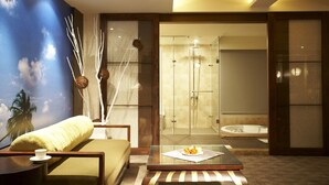 Una habitación de hotel moderna con una mesa de centro de vidrio, un sofá, una pared decorativa con un mural tropical y una zona de ducha separada con paredes de vidrio.