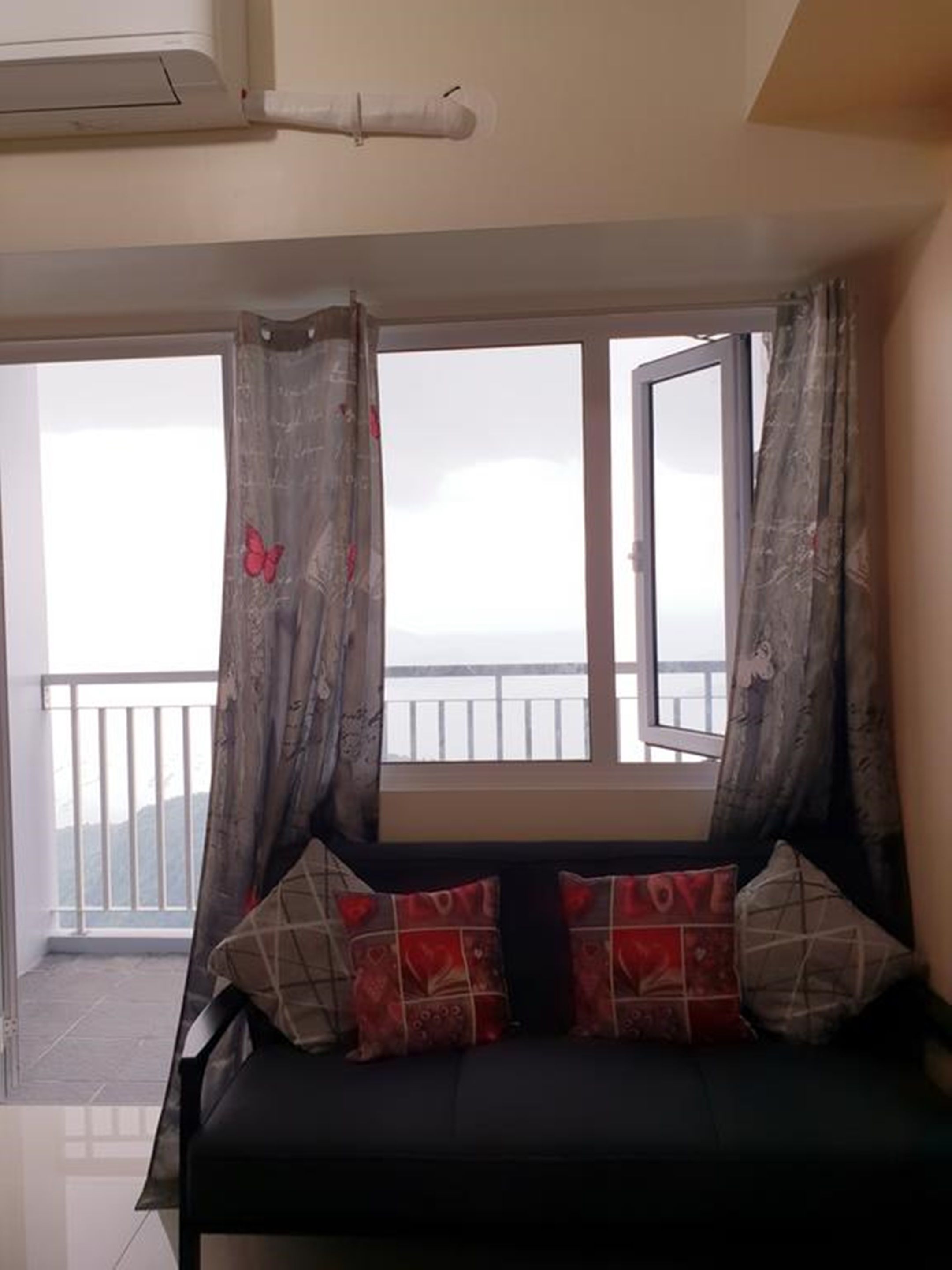 Una habitación con un sofá negro, cojines estampados y vistas a un balcón con barandilla.