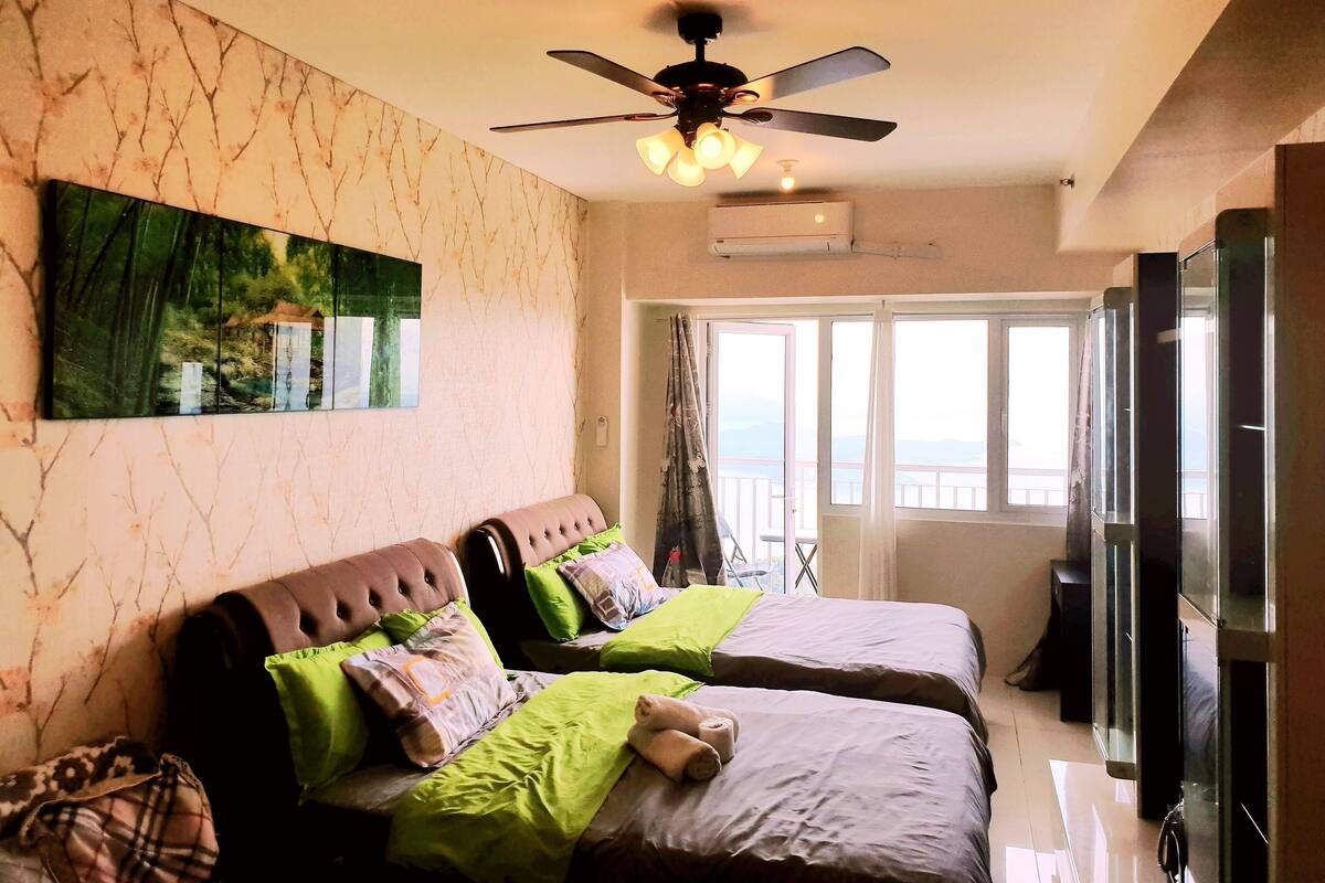 A hotel room with two beds, a ceiling fan, a large window with a view, and a painting on the wall.