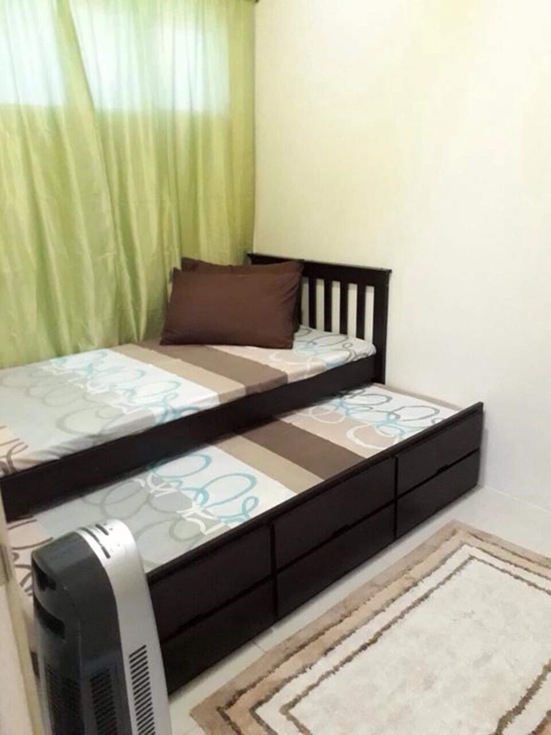 A small, single-bed room with a black frame, a patterned bedspread, a brown pillow, and a bedside table with a lamp.