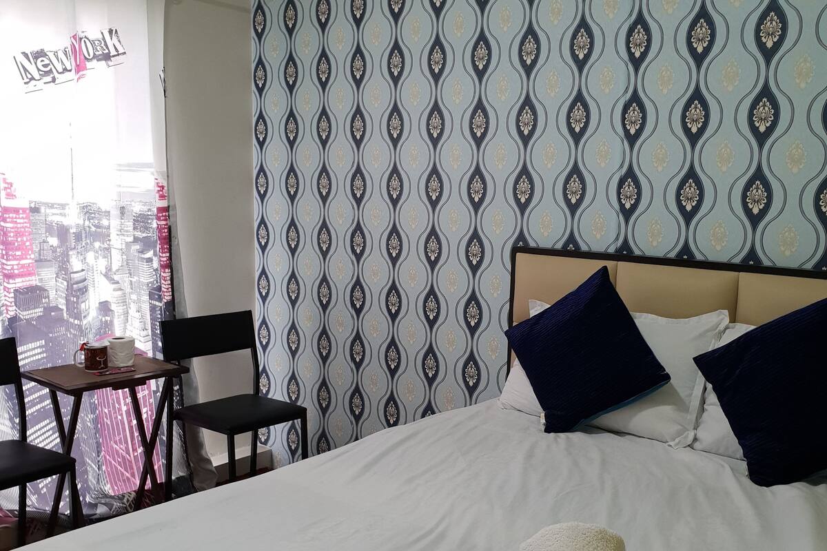 A hotel room with a bed, two pillows, a table with a chair, and a view of the cityscape through a window.