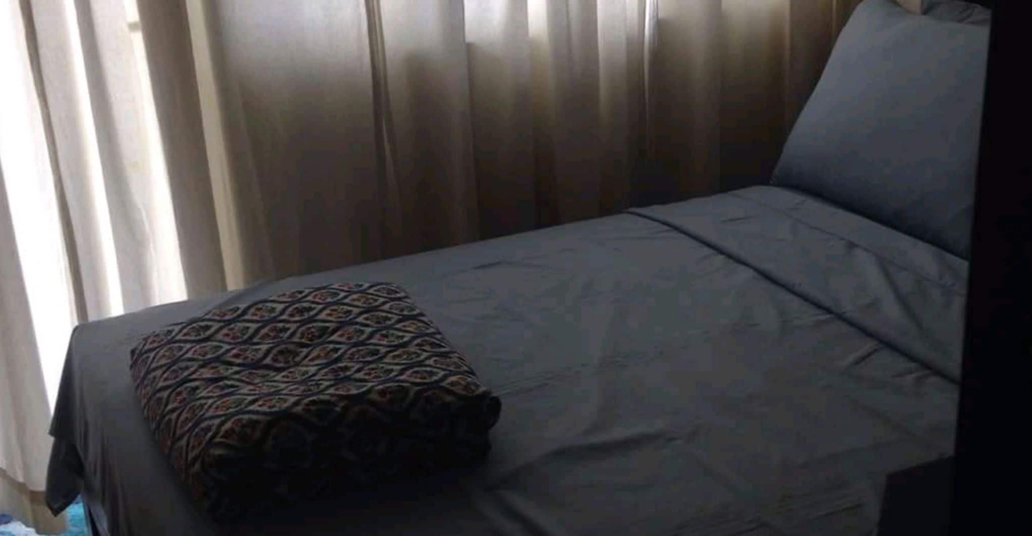 A bed with a patterned pillow and a plain grey bedspread in a room with vertical blinds.