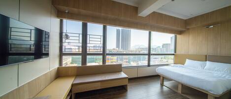 A modern hotel room with a large window, a bed, a bench, and a TV.