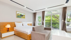 Habitació d'hotel moderna amb un llit de fusta, un sofà beix, un escriptori blanc i grans finestres amb cortines.