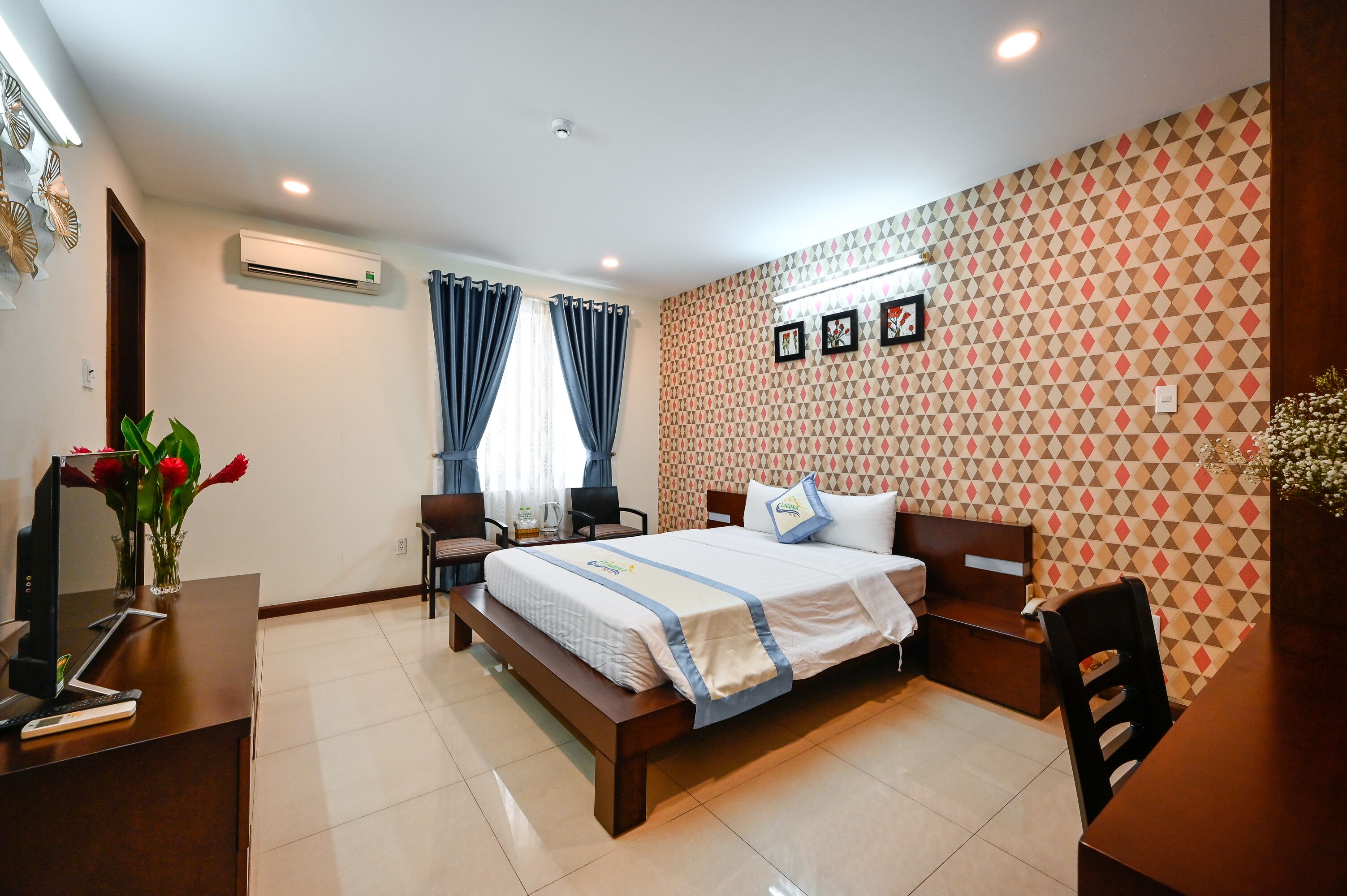 A hotel room with a bed, desk, and chair, a desk lamp, and a vase with flowers.