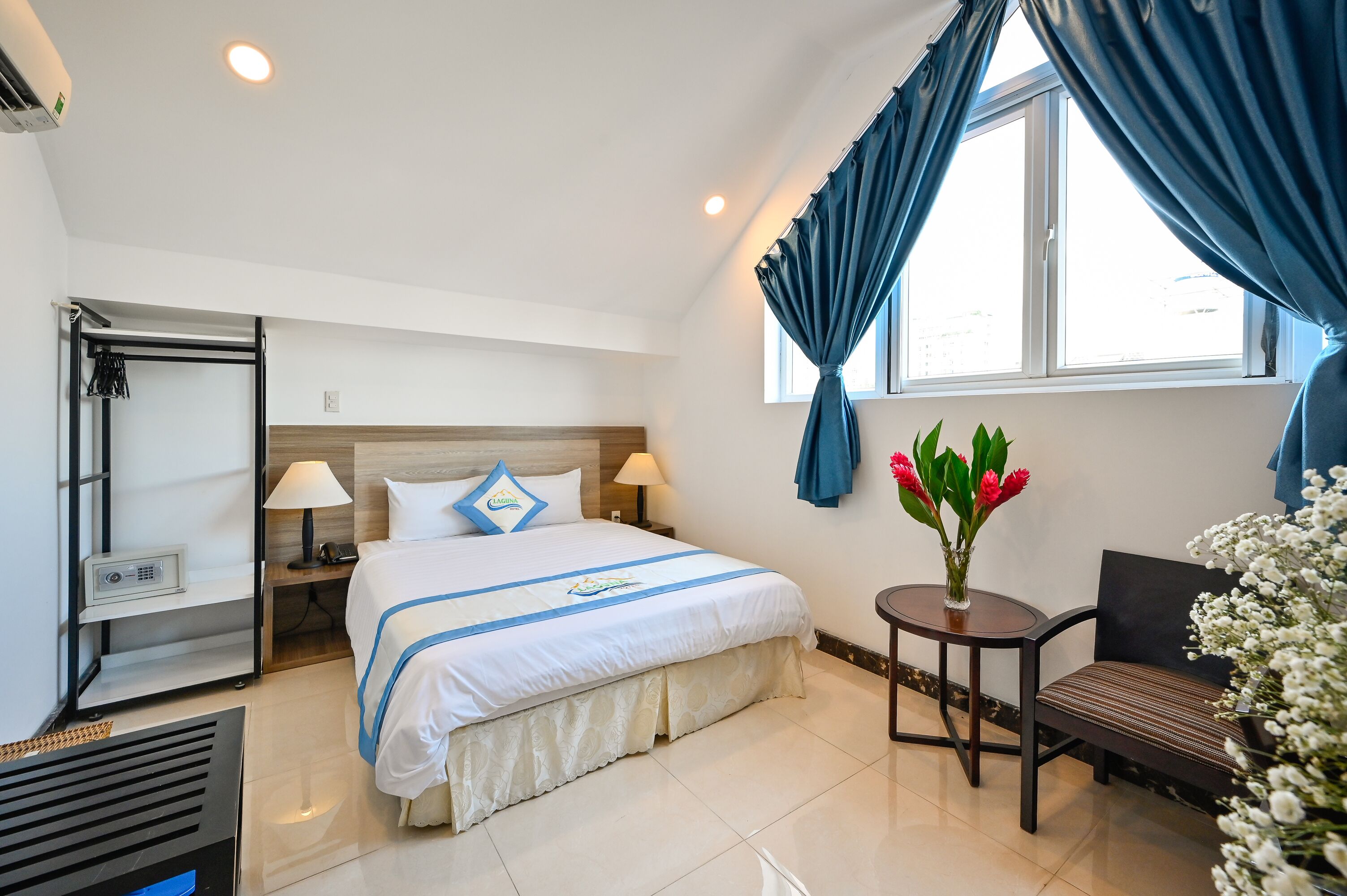 A hotel room with a bed, bedside lamps, a small table with flowers, a chair, and a window with curtains.