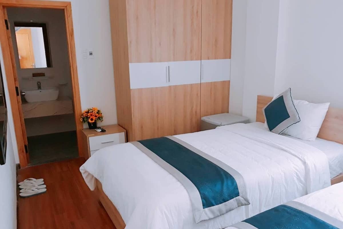 A hotel room with two beds, a wooden wardrobe, a bedside table with a lamp, a small table with a vase of flowers, and a bathroom visible through an open door.