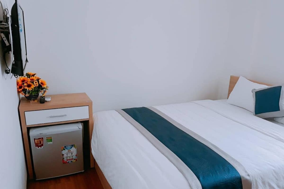 A hotel room with a bed, a small desk, a refrigerator, and a vase with flowers.